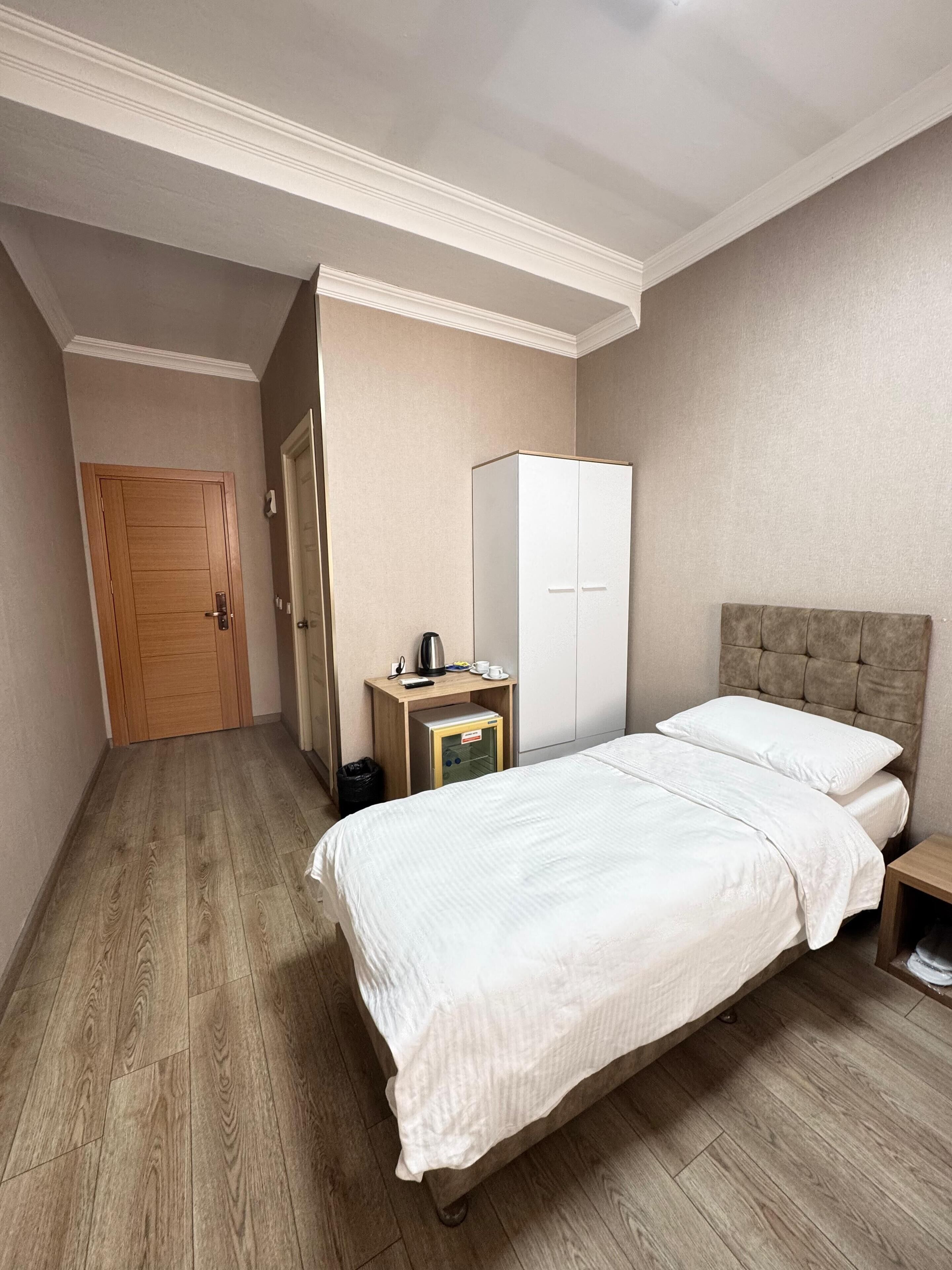 Quarto de hotel com piso de madeira, cama com roupa de cama branca, guarda-roupa, mesa de cabeceira com abajur e uma porta.