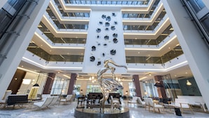 A modern hotel lobby with a central sculpture, multiple floors, and a seating area with tables and chairs. - Flora Garden Ephesus Hotel Kusadasiot (Kusadasi)