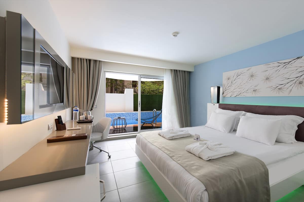 A hotel room with a bed, a TV, a desk with a chair, a window with curtains, and a view of an outdoor pool.