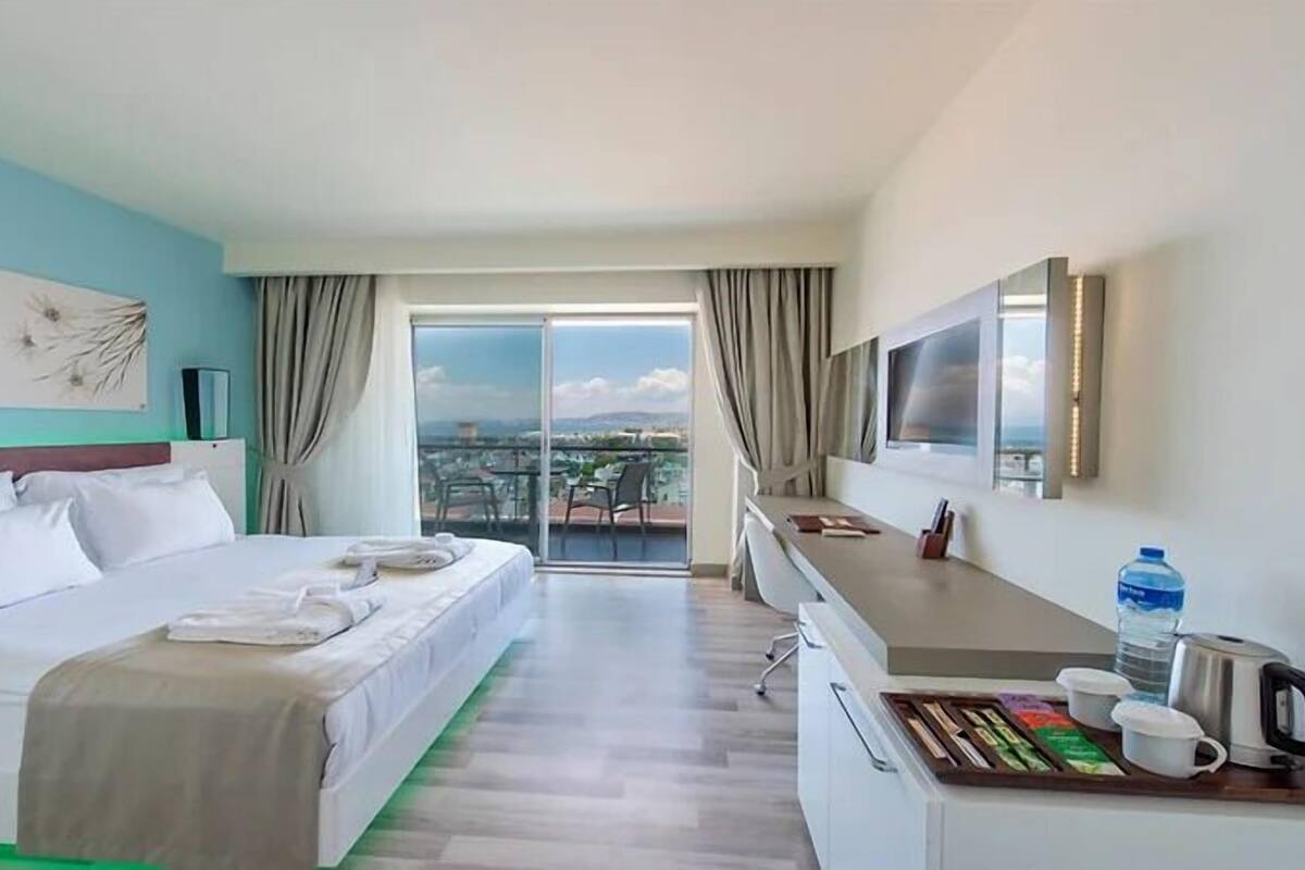 A hotel room with a bed, a desk with a coffee maker, a TV, and a view of the city through the balcony.