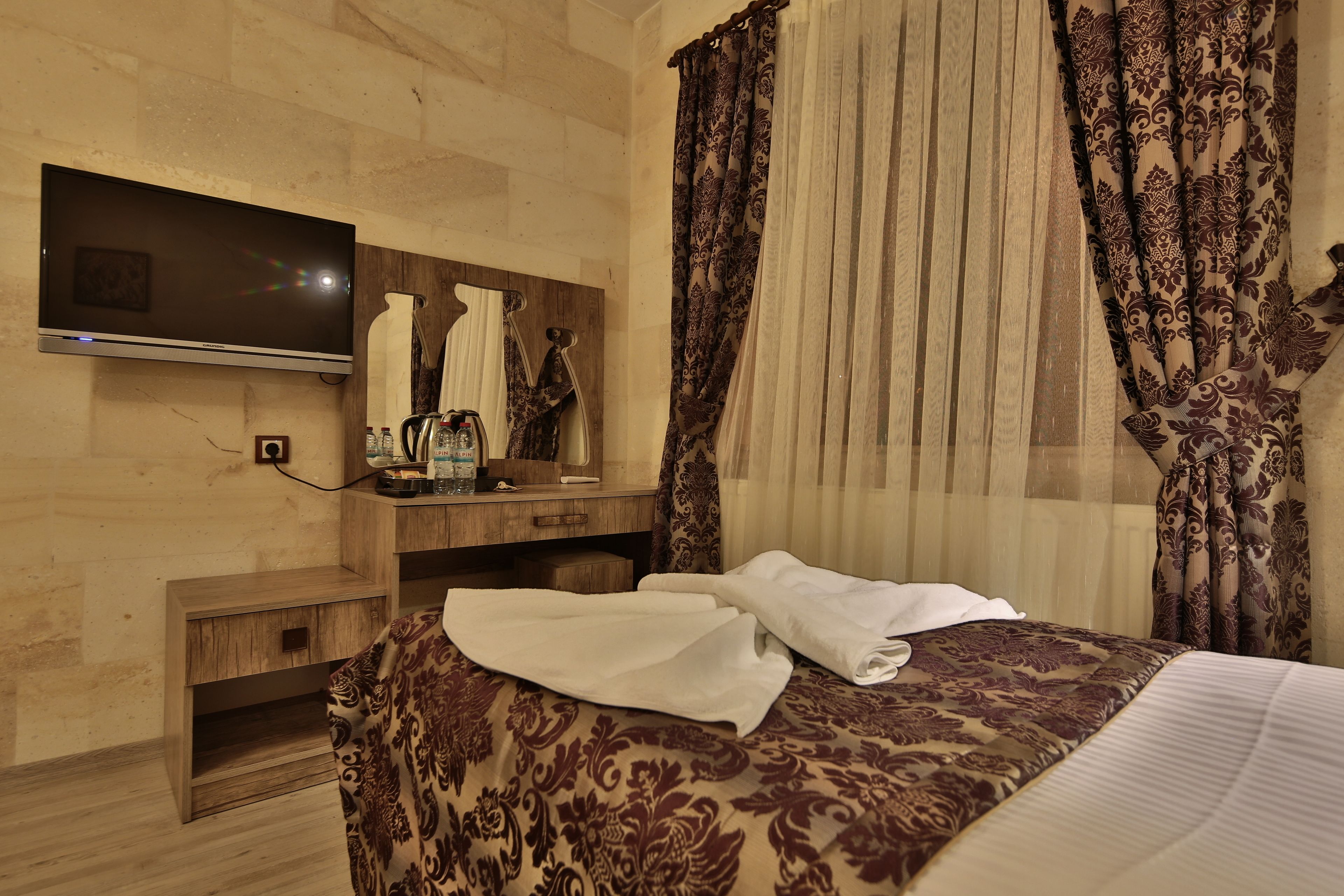 A hotel room with a bed, a TV mounted on the wall, a wooden nightstand, and a mirror.