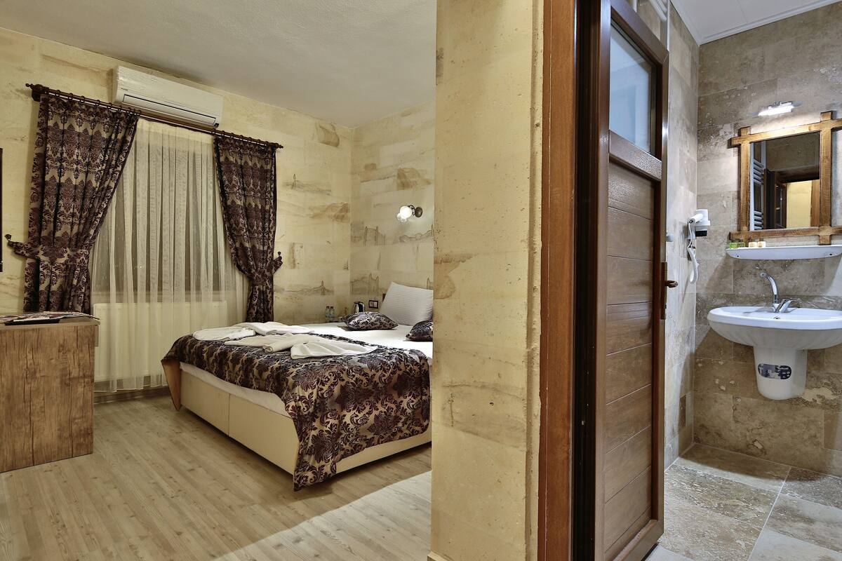 Une chambre à coucher avec un lit, des rideaux, une table de chevet et une salle de bain équipée d’un lavabo et d’un miroir.
