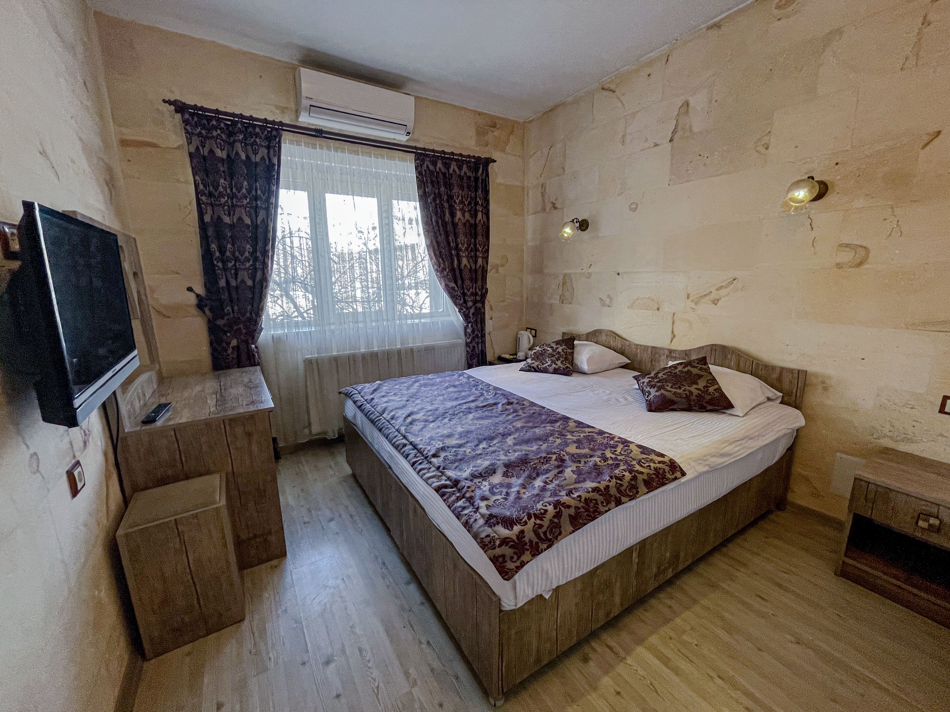 Un dormitorio con cama de madera, televisor sobre un mueble bajo, ventana con cortinas y equipo de aire acondicionado.