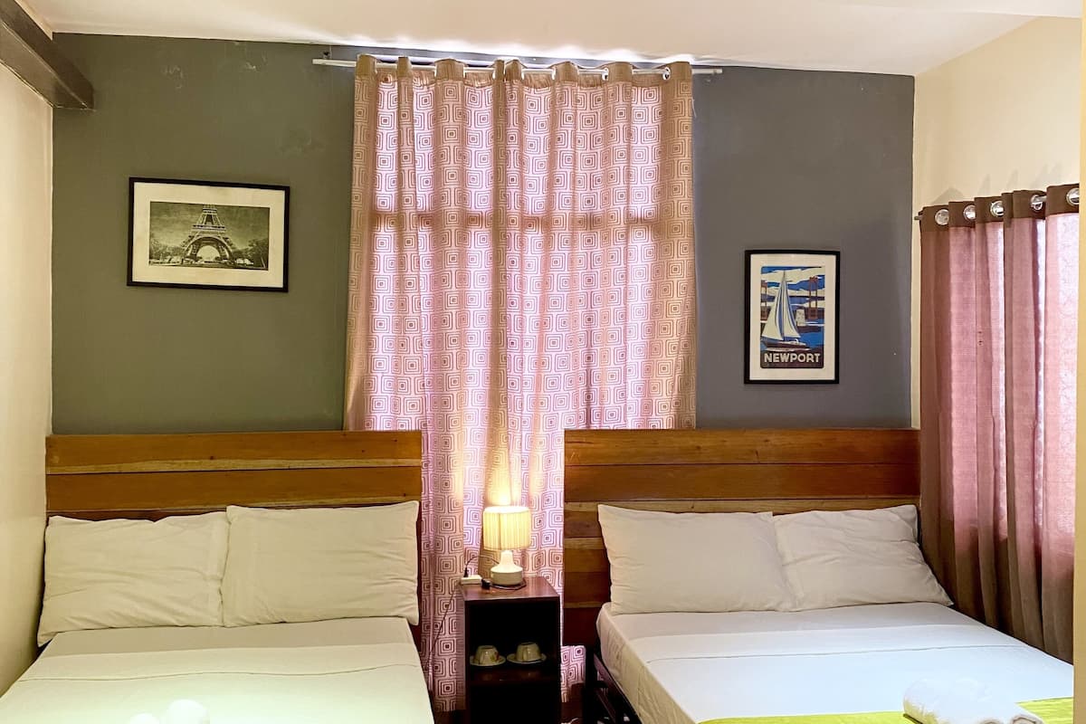 A hotel room with two single beds, wooden headboards, a nightstand with a lamp, and a window with a curtain.