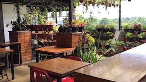 An indoor dining area with wooden tables and red chairs, surrounded by potted plants and a view of greenery outside. - Aliria Bed and Breakfast (Tagbilaran)