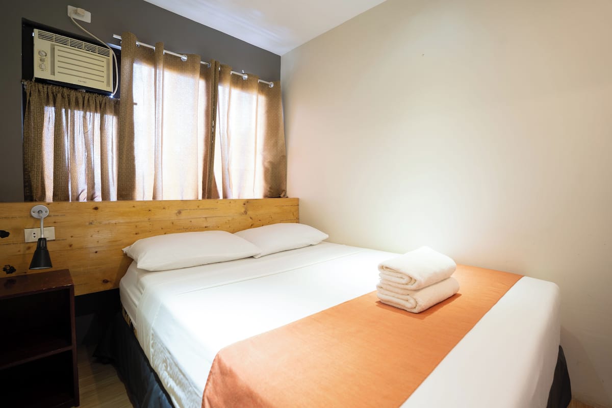 A hotel room with a wooden headboard, a bed with white linens, a wooden nightstand, and a window with curtains.