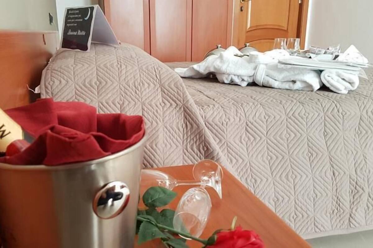 A hotel room with a bed, a minibar, a tray with a champagne bottle, a glass, and a rose.