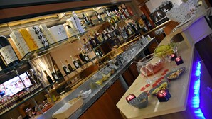 A bar area with shelves of bottles and glasses, a counter with food and drinks, and a television screen in the background. - Hotel Joy (Castel Volturno)
