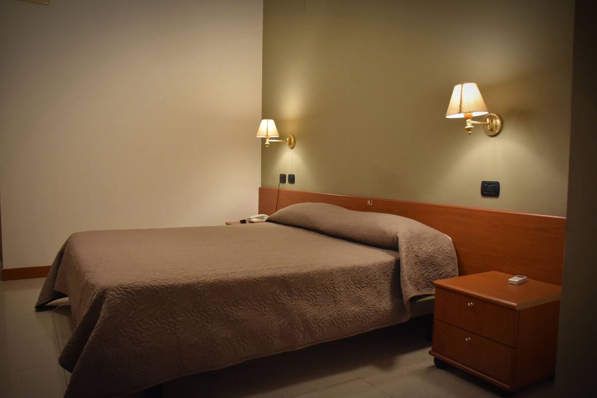 A neatly made bed with a brown bedspread, two wall-mounted lamps, and a wooden nightstand with a drawer.