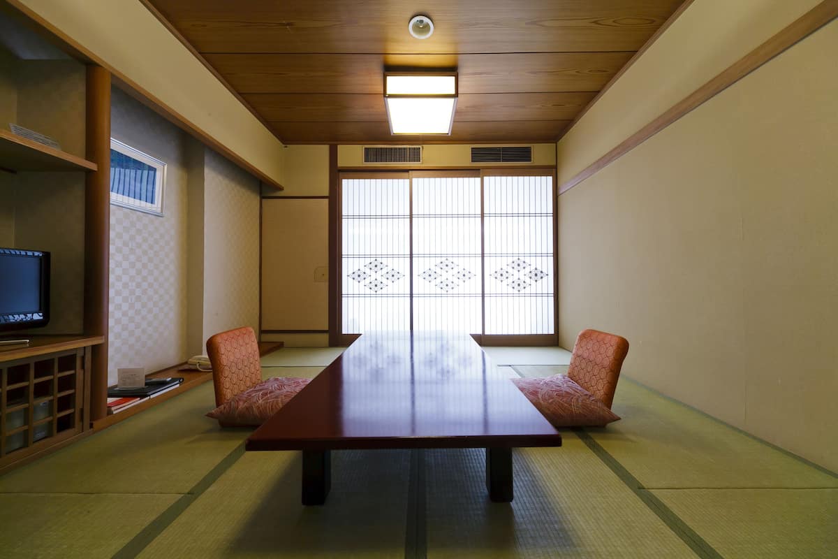 A traditional Japanese room with tatami flooring, a low wooden table, two tatami mats with cushions, a television on a shelf, and shoji screens.