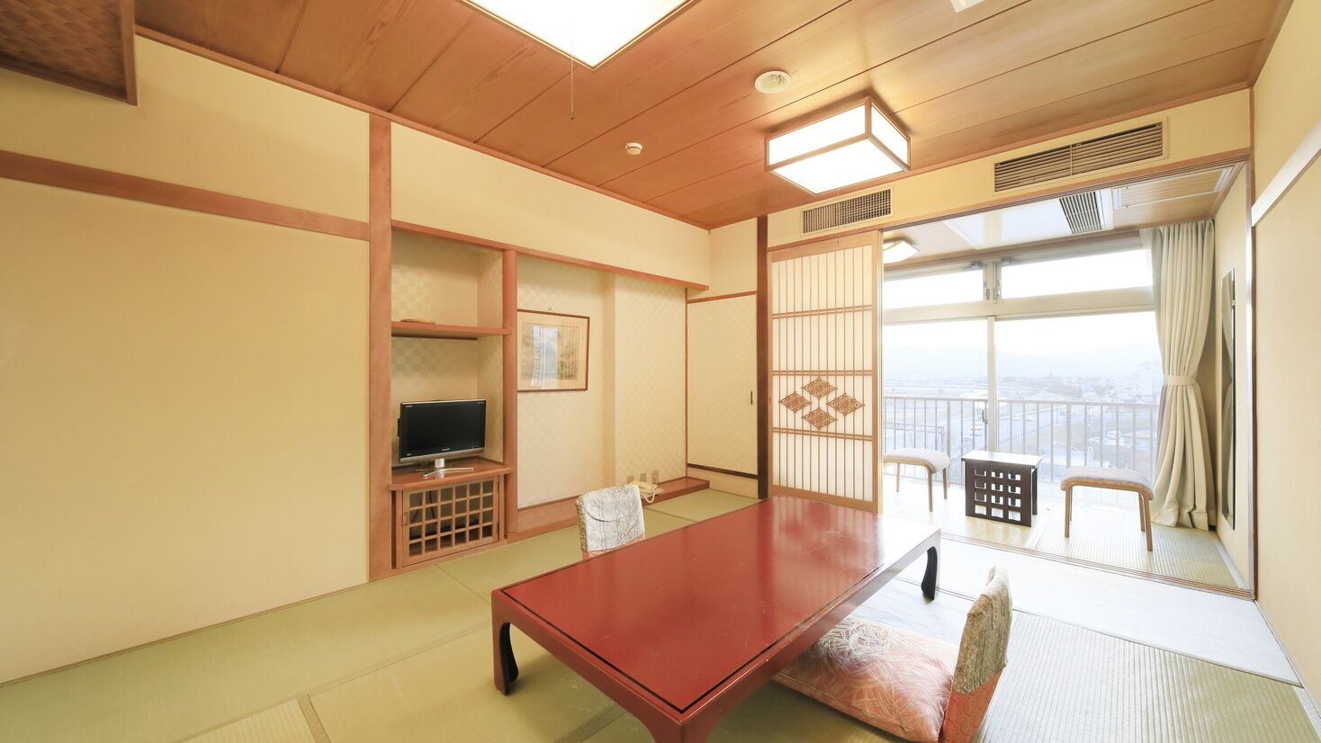 A traditional Japanese room with tatami flooring, a low wooden table, a television on a stand, and a sliding door leading to a balcony with a view.