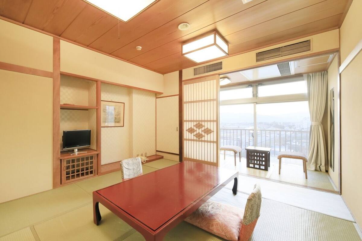 A traditional Japanese room with tatami flooring, a low wooden table, a television on a stand, and a sliding door leading to a balcony with a view.