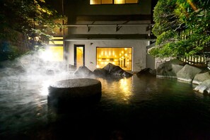 A traditional Japanese onsen with a stone bath in the foreground, a building with a wooden door, and a view of the surrounding trees and rocks. - Fujinoya Yutei (Fuefuki)
