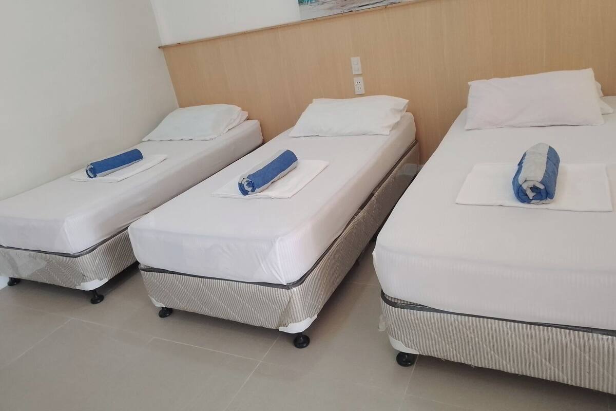 Three single beds with white linens and blue towels, a wooden headboard, and a painting on the wall.