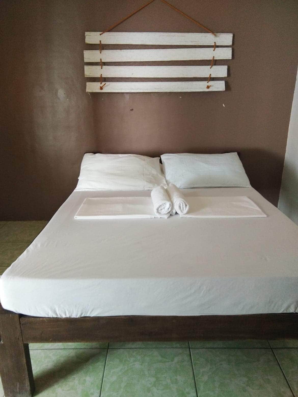 Un lit bien fait, avec du linge de lit blanc et une serviette enroulée, une tête de lit en bois et un mur de couleur marron.
