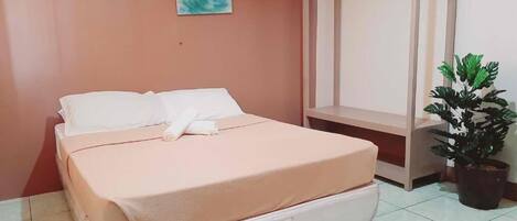 Ein Einzelbett mit weißer Matratze und rosa Bettdecke, ein Nachttisch, ein Topf mit Pflanze und ein an der Wand befestigtes Kunstwerk.