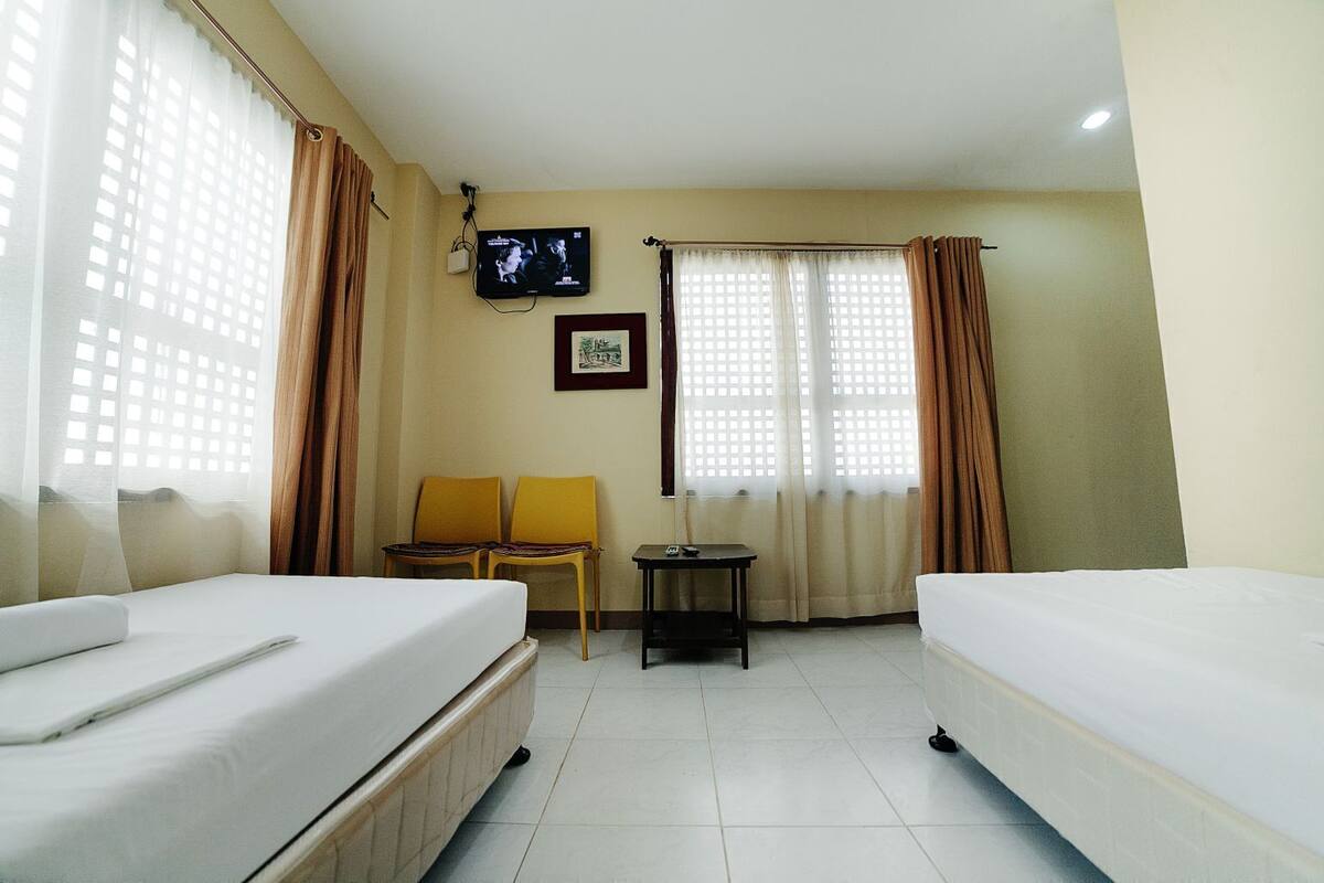 A hotel room with two beds, a TV, a small table, two chairs, and large windows with curtains.