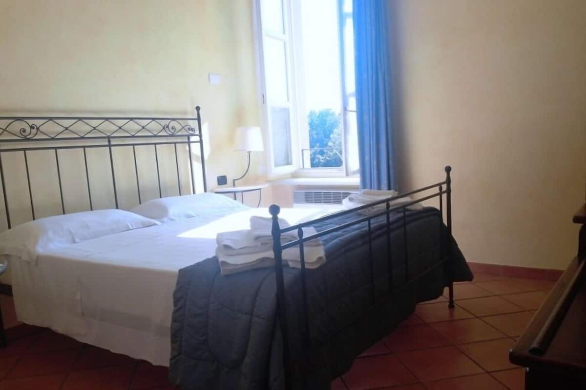 A single bed with a metal headboard, a window with curtains, and a tiled floor.