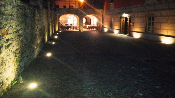 Une rue pavée menant à un bâtiment doté d’arcades et dont l’intérieur est éclairé, ce qui évoque un cadre historique ou traditionnel.
