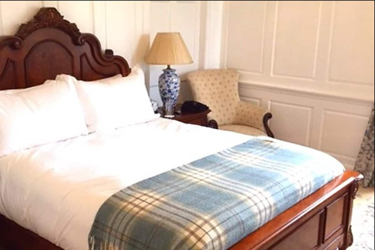 Una cama bien hecha con una manta escocesa, un cabecero de madera, una mesita de noche con lámpara y un sillón con estampado.