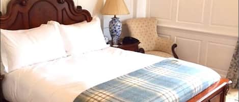A neatly made bed with a plaid blanket, a wooden headboard, a bedside table with a lamp, and a patterned armchair.