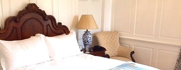 Une chambre à coucher avec une tête de lit en bois, une table de chevet avec une lampe, un fauteuil à motifs et une porte blanche en arrière-plan.