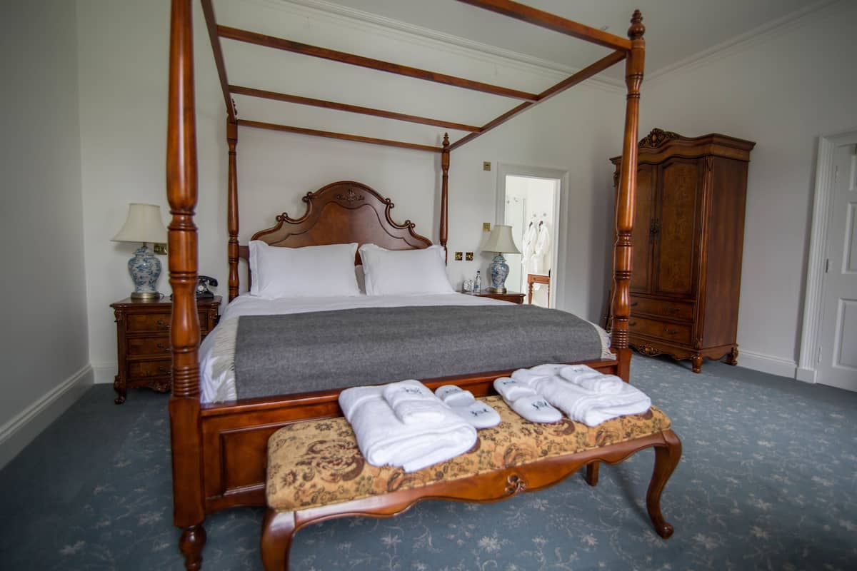 Una cama con dosel y tablero de madera, un banco con toallas y una mesita de noche con lámpara.