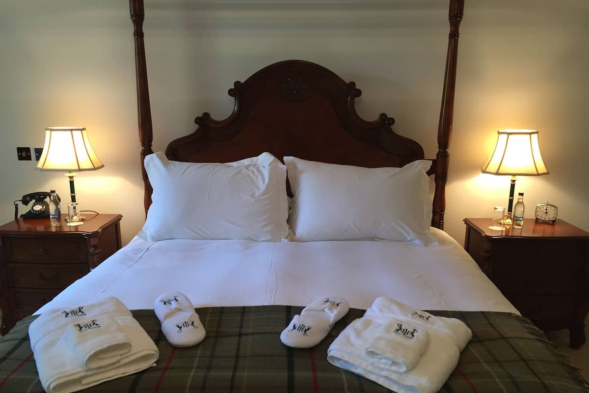 Una cama bien hecha con sábanas blancas, dos almohadas y una colcha de cuadros. Hay dos mesitas de noche con lámparas, un reloj y una botella en una.