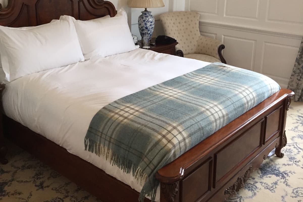 Una cama bien hecha con una manta escocesa, un cabecero de madera, una mesita de noche con una lámpara y una silla.