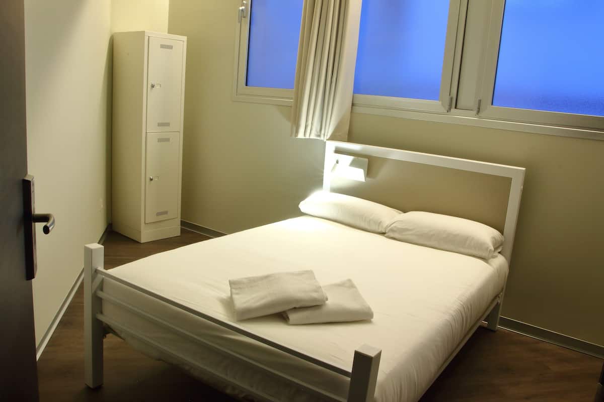 Un lit bien fait, avec du linge de lit blanc et deux serviettes pliées, une armoire blanche et une fenêtre avec des rideaux.