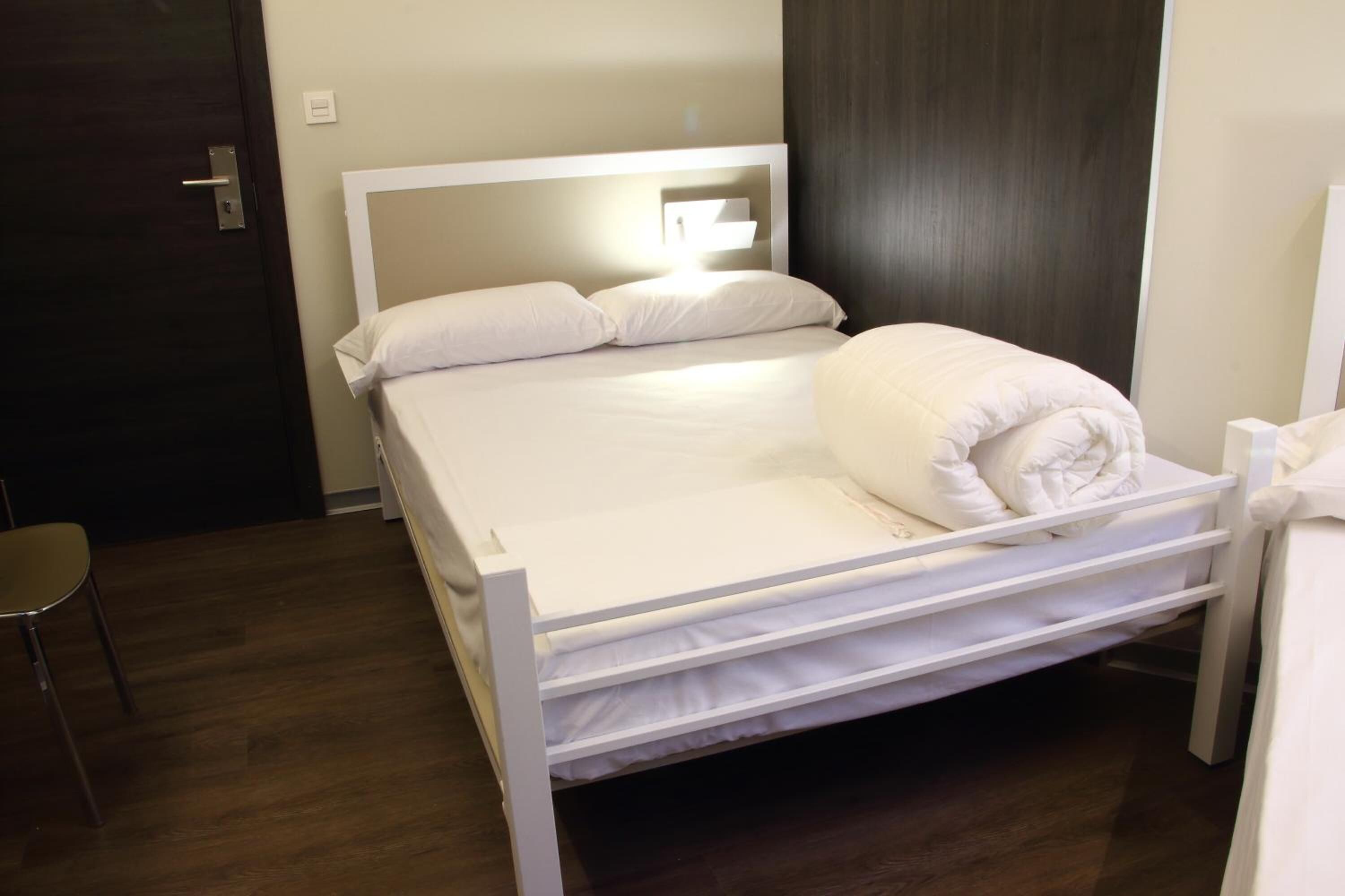 Una cama individual con cabecera y piecera blancos, una mesita de noche y una silla.