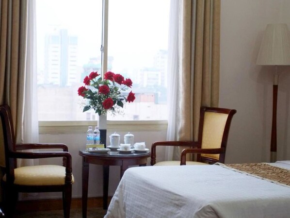 A hotel room with a bed, two chairs, a small table with a vase of red flowers, and a window with a view of buildings. - Vietnam Trade Union in Hanoi (Hanoi)