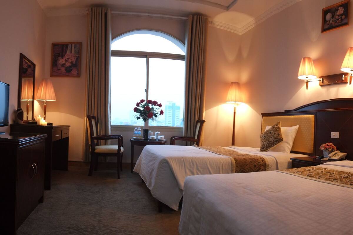 A hotel room with two beds, a desk, a chair, a television, and a window with curtains.