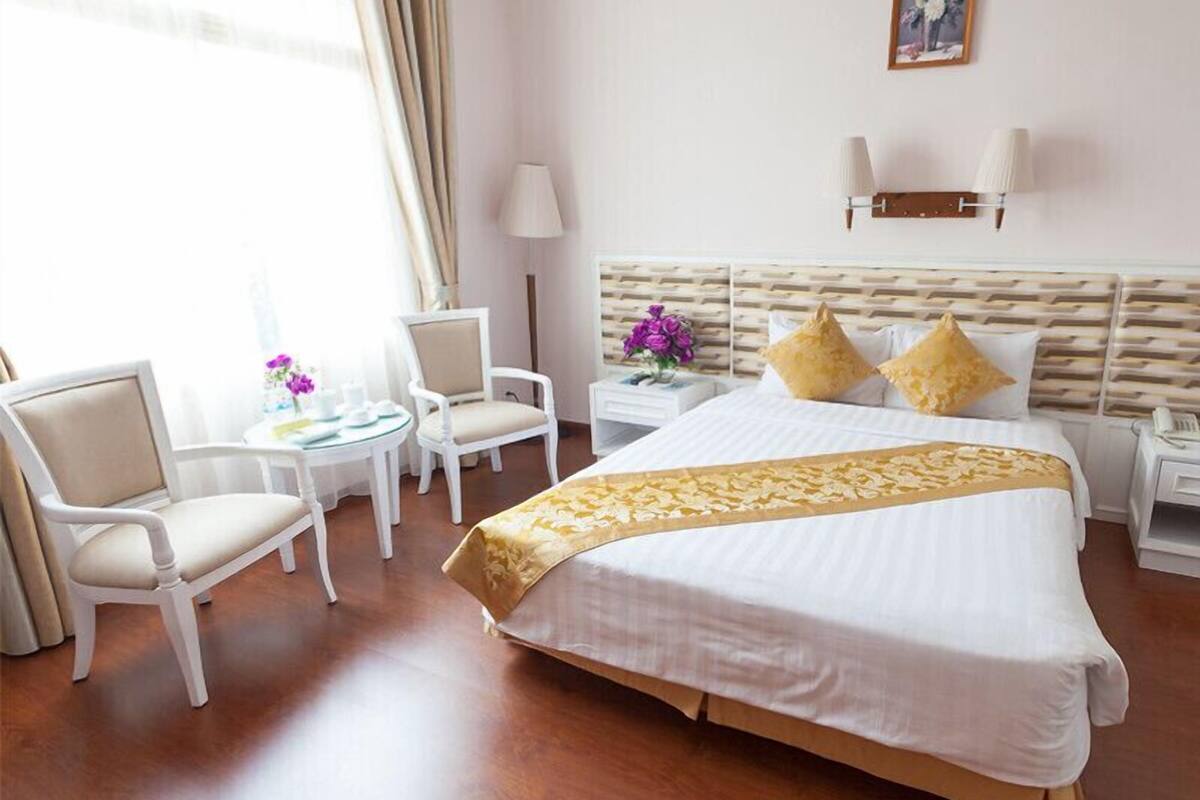 A hotel room with a large bed, two chairs, a small table, a nightstand, and a wall-mounted lamp.