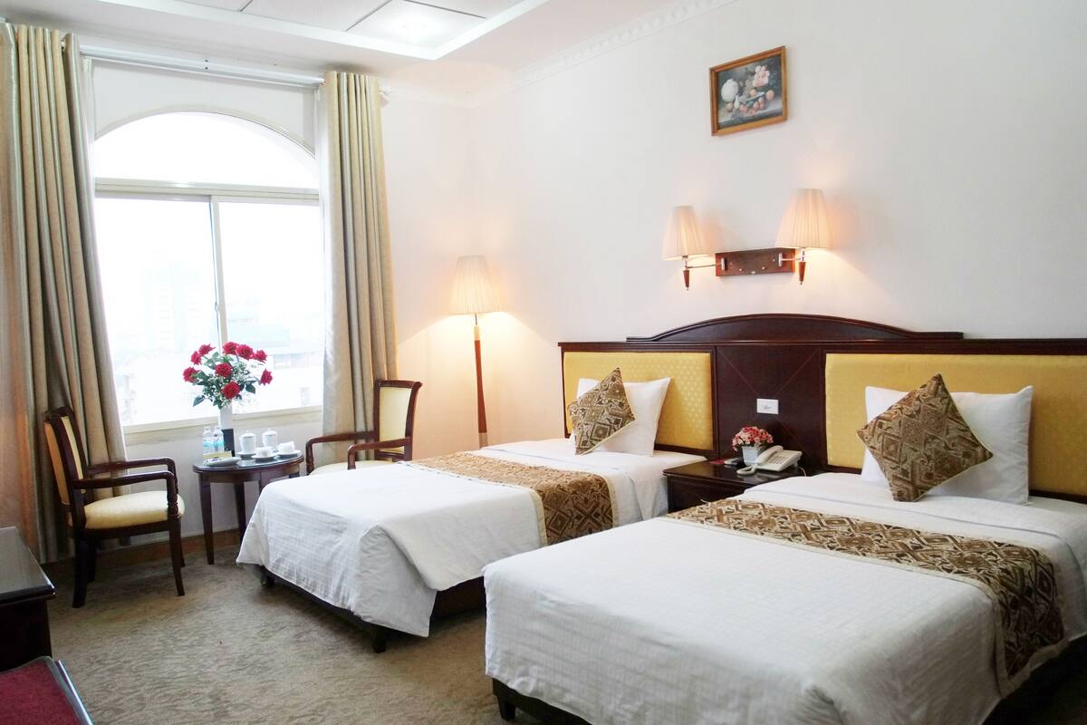 A hotel room with two beds, a chair, a small table with a vase of flowers, and a window with curtains.