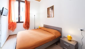 Een slaapkamer met een bed, nachtkastje, lamp, tv die aan de muur is bevestigd en een raam met gordijnen.