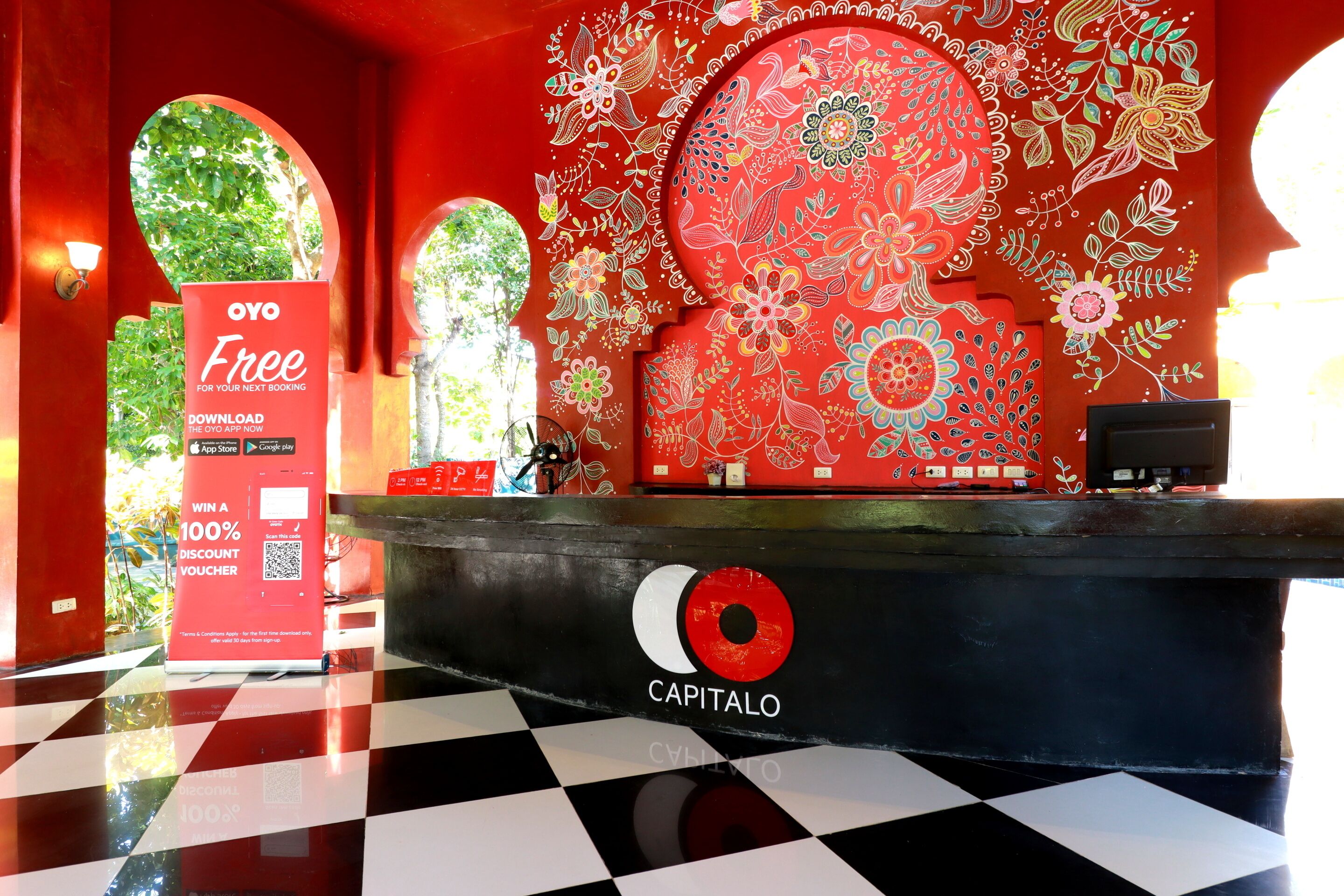 Zona de recepción de un hotel con piso a cuadros, diseño floral rojo y blanco en la pared y el logo 'CAPITALO' en el mostrador.