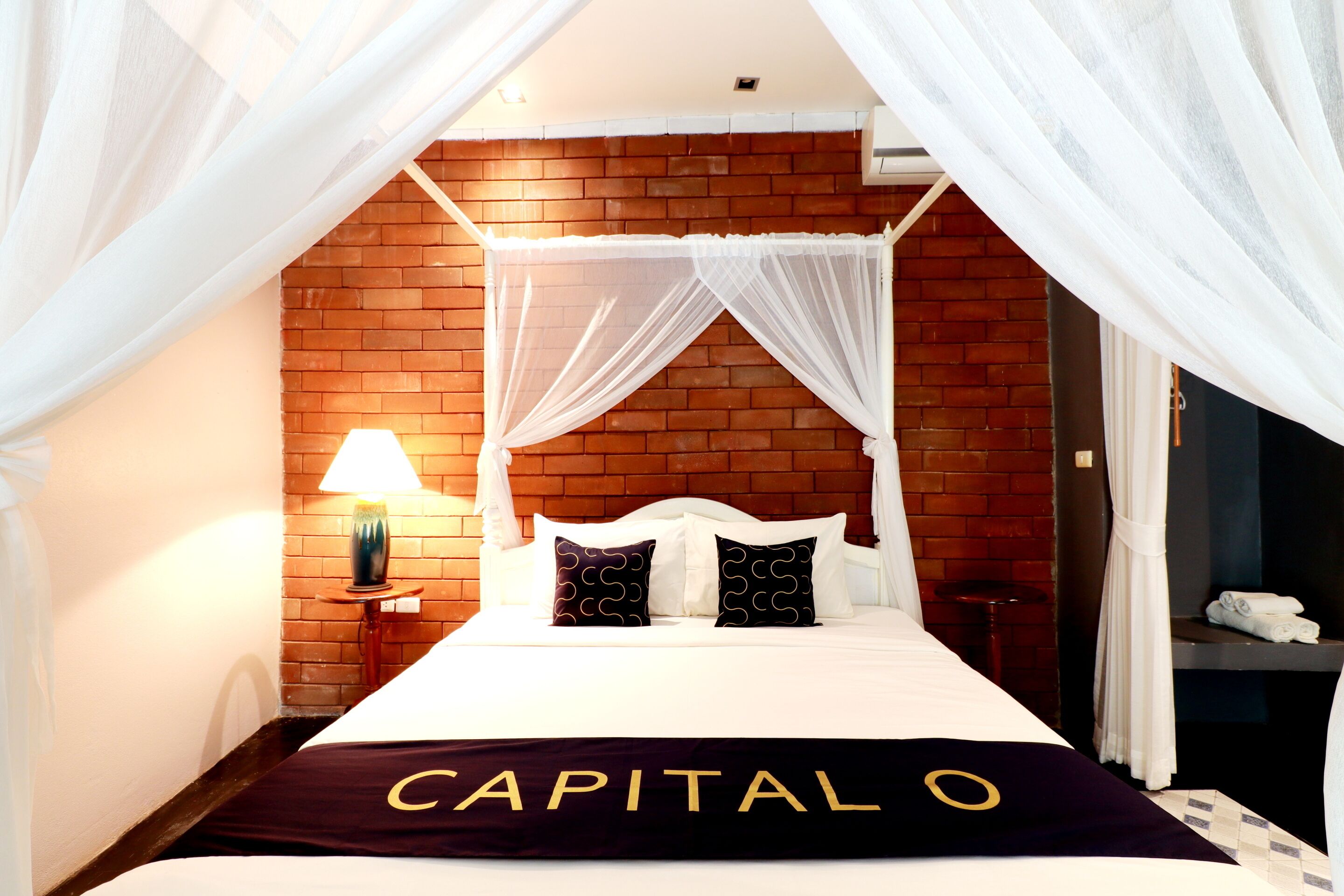 A bed with a canopy, a brick wall, a lamp, and a towel rack.