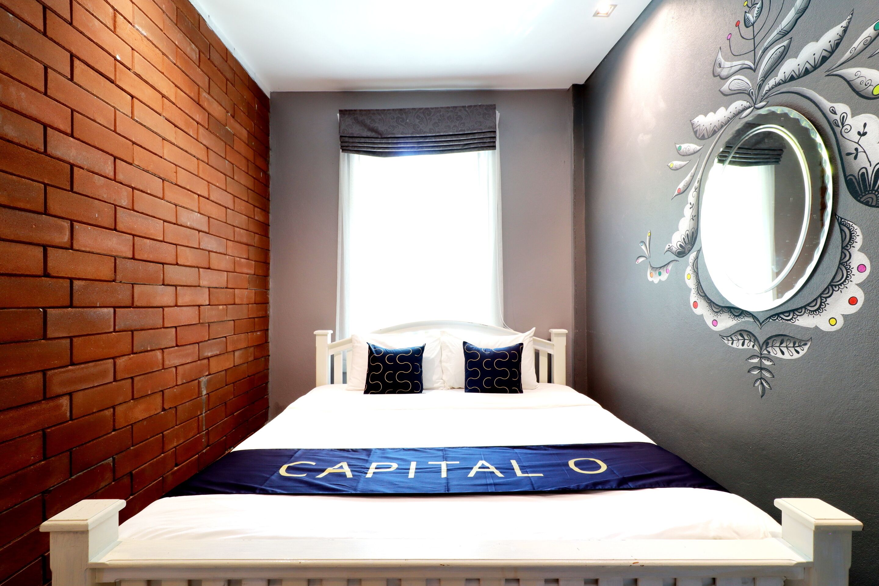 A bed with a white frame, a blue and white bedspread, and a pillow with a circular pattern. A window with a dark valance is visible in the background.