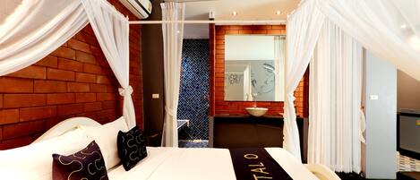 A hotel room with a bed, white curtains, a brick wall, a bathroom with a sink, and a mirror.