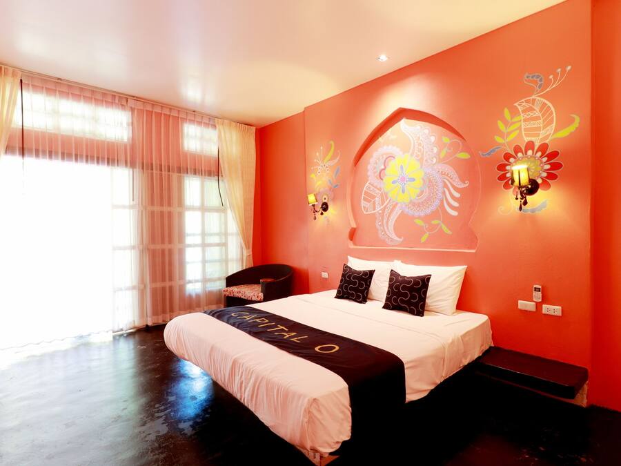 A hotel room with a large bed, a desk, and a chair. There is a window with curtains, a wall-mounted light, and a decorative wall piece.