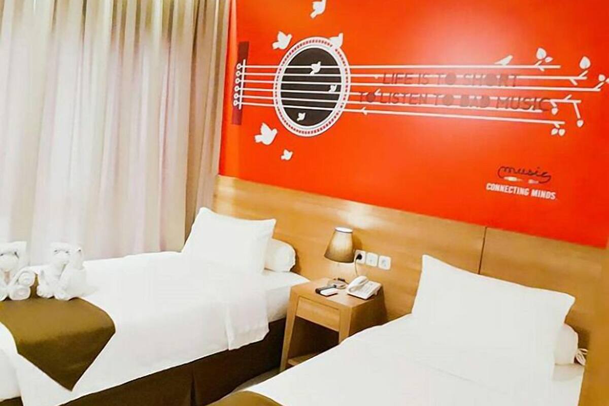 Habitació d'hotel amb dos llits, un capçal de llit de fusta, una meseta de nit amb una làmpada, i una paret amb un disseny vermell que inclou notes musicals i el text "FRONTONE HOTEL".