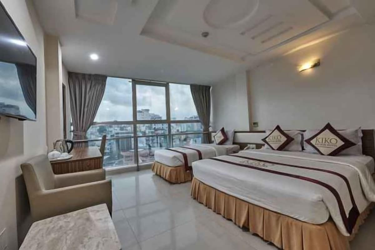 Habitación de hotel con dos camas, un sofá, un televisor y un amplio ventanal con vistas a la ciudad.