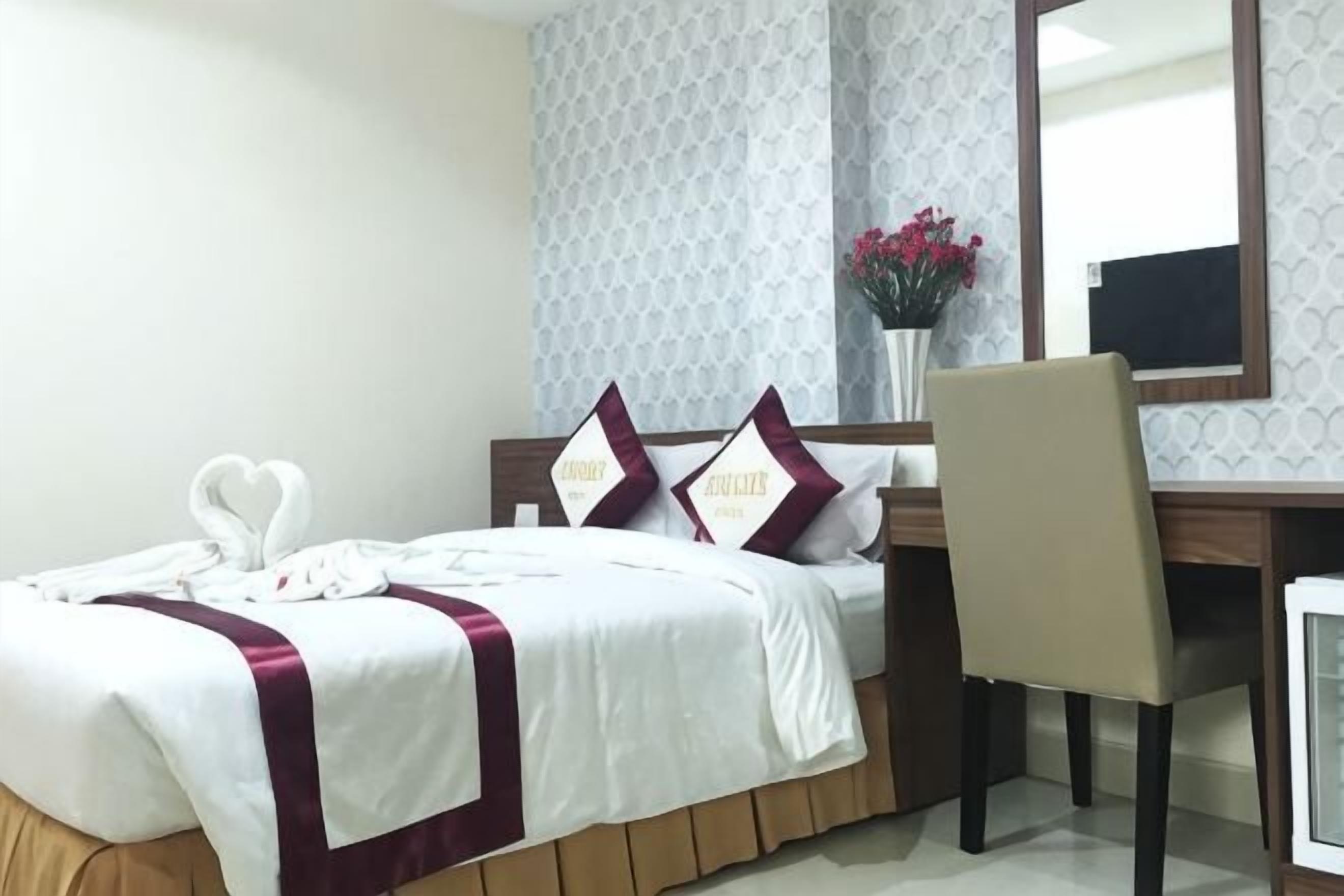Une chambre d’hôtel avec un lit, un bureau, une chaise, un miroir et un vase avec des fleurs.