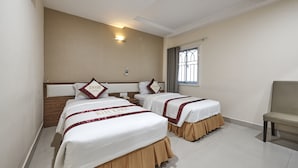 Habitación de hotel con dos camas, cada una con cabecera y almohada decorativa. Se observa una ventana con cortinas, una silla y una lámpara fijada a la pared.