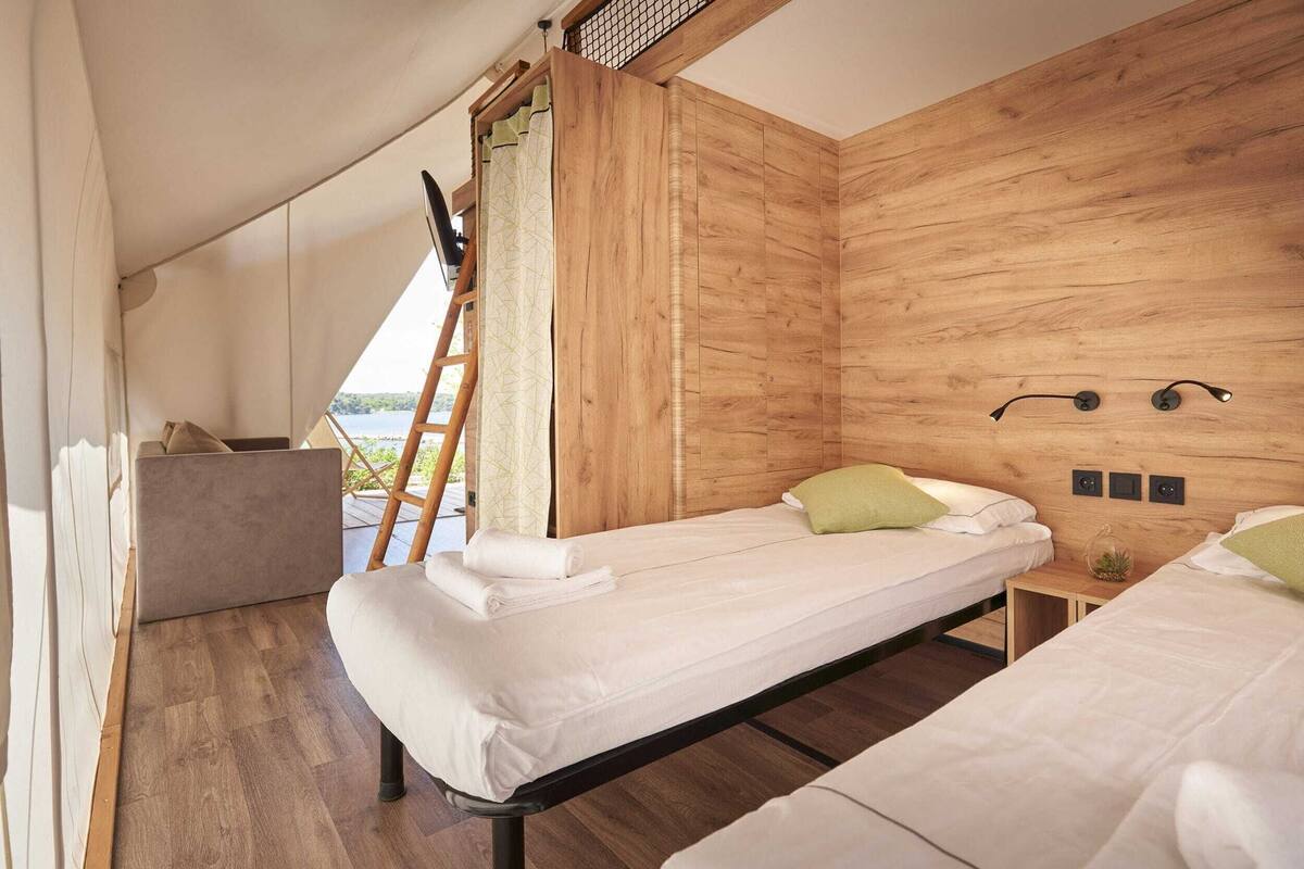 A room with two beds, a wooden wall, a small table, and a view of the outdoors.