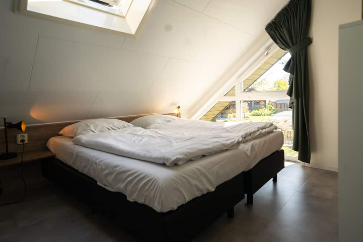 A bedroom with a skylight, a bed, a bedside table with a lamp, and a view of the outside.