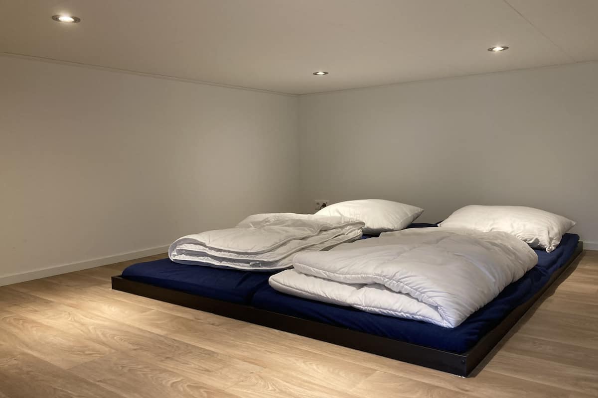 A single bed with a blue mattress in a room with wooden flooring and recessed ceiling lights.
