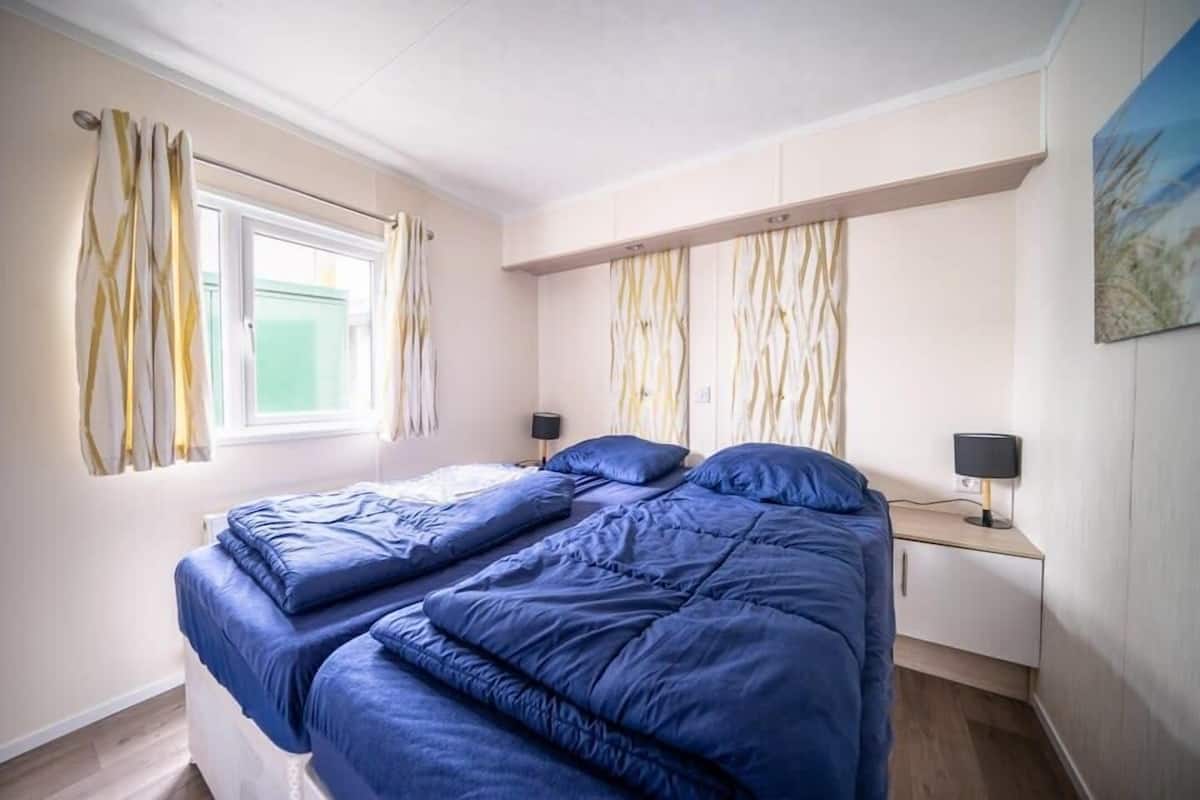 A bedroom with two beds, a window with curtains, a bedside table with a lamp, and a painting on the wall.
