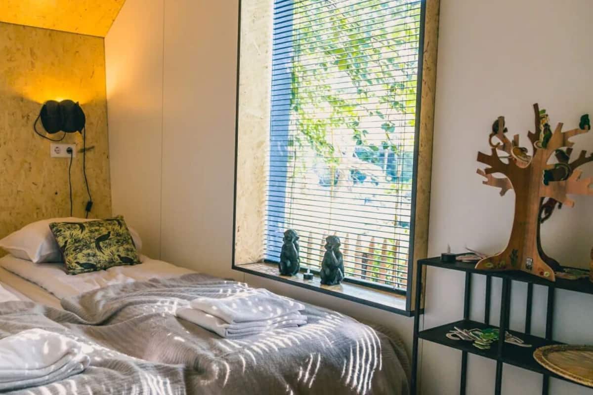 A room with two beds, a window with blinds, a wall-mounted shelf with decorative items, and a wooden headboard.